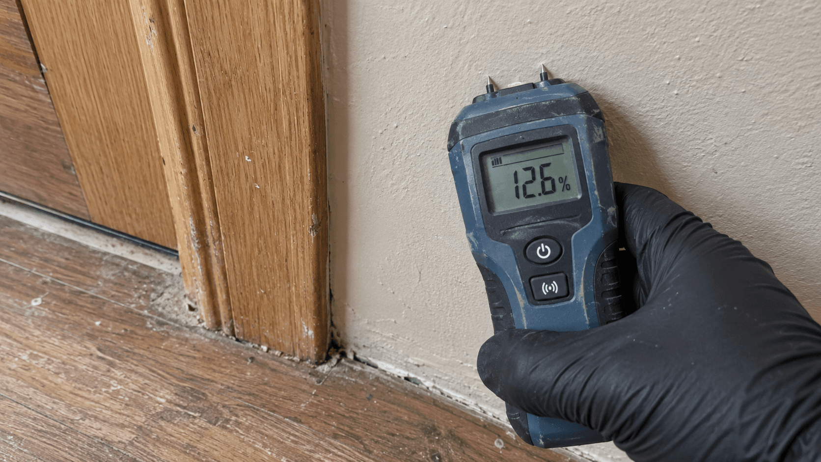 Gloved hand checking a wall with a moisture meter beside a wood door frame